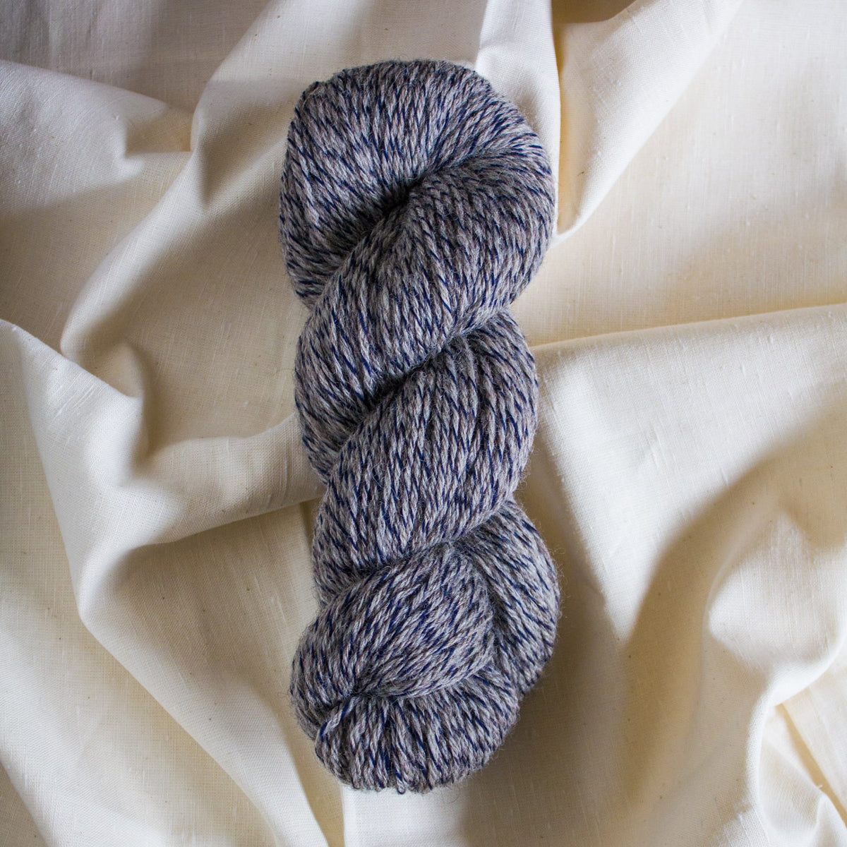 Full skein of Garthenor Organic Unruly: Folly yarn, a DK weight limited-edition blend of British Romney, Polwarth, and Hebridean wool in a grey and navy marl, shown on undyed natural fabric.